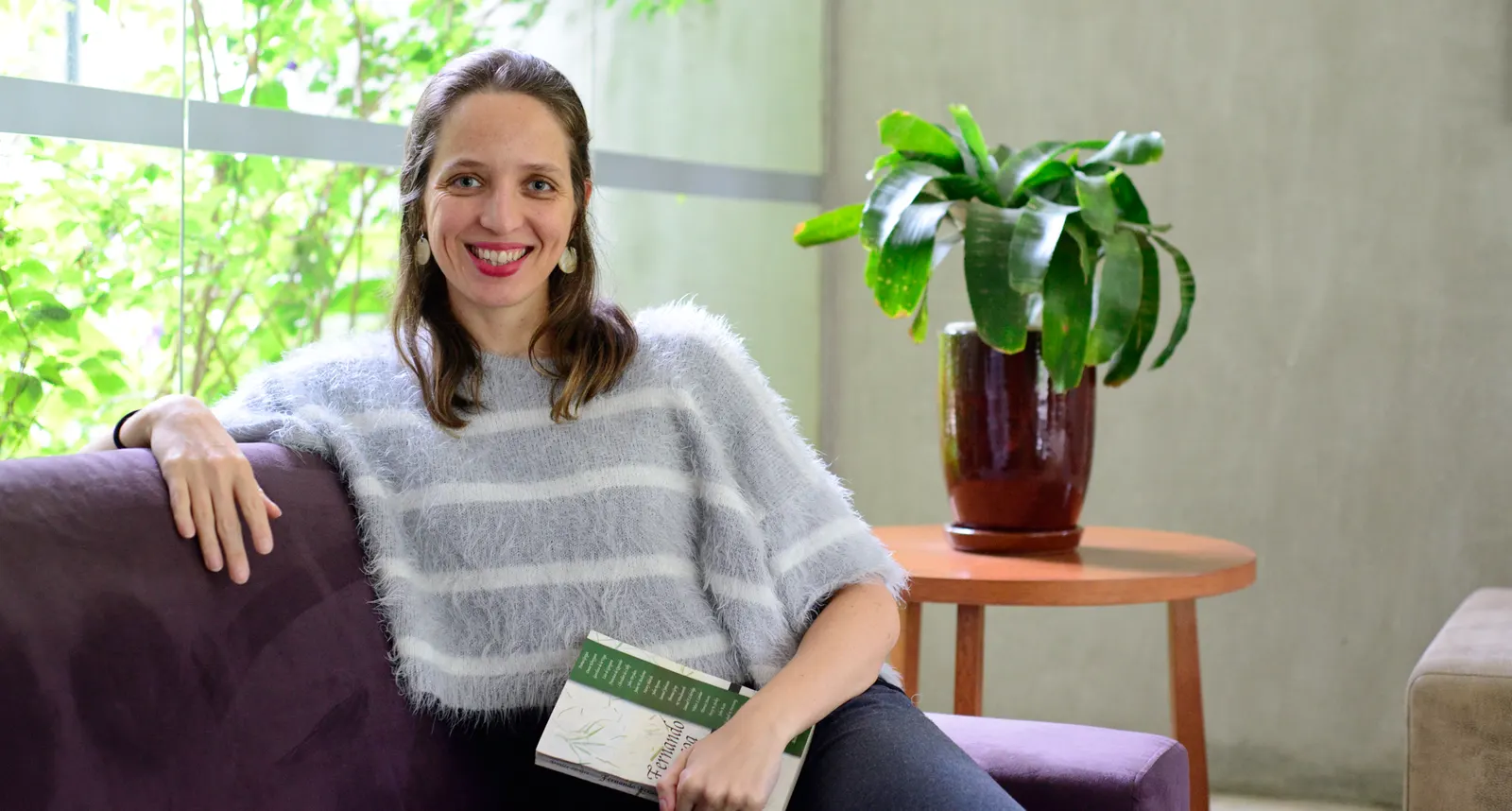 Mirella Duarte sentada em um sofá com um livro nas mãos, ao lado de uma planta e de uma janela com vegetação